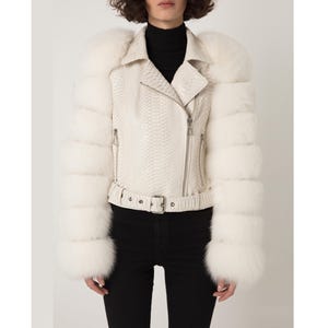 Può includere: Giacca in pelle color crema con motivo a pelle di serpente, con colletto a revers, cerniera diagonale e cintura in vita. Le maniche sono decorate con pelliccia bianca. La giacca è indossata sopra un dolcevita nero.