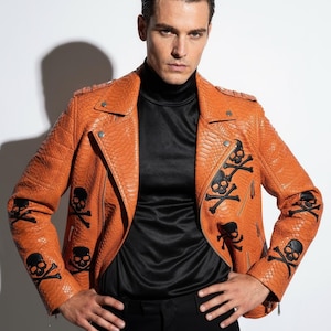Peut inclure: Veste en cuir orange avec une texture peau de serpent et des appliqués de têtes de mort et d'os croisés noirs. La veste a un style motard classique avec un col cranté et une fermeture éclair. Le modèle porte un col roulé noir et un pantalon noir.