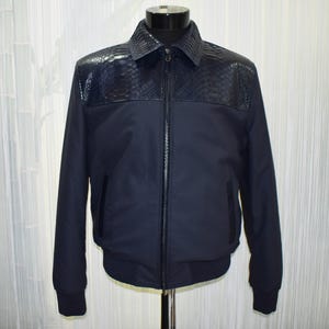 May include: A navy blue jacket with a textured, dark blue panel across the shoulders and collar. The jacket has a zip-up front, side pockets, and ribbed cuffs and hem. The jacket is displayed on a mannequin.