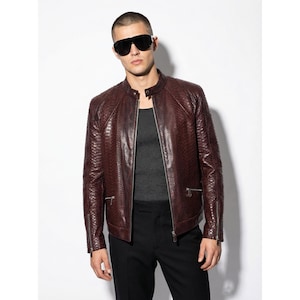 以下が含まれることがあります： テクスチャード加工が施されたバーガンディ色のレザージャケット。 ジャケットはジッパーフロント、サイドポケット、スタンドカラーが特徴です。 画像の人物は黒いサングラス、グレーのタンクトップ、黒いパンツを着用しています。