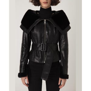 May include: A black leather jacket with a snakeskin pattern, a fur collar, and fur cuffs. The jacket has a belt with a rectangular buckle and a zip closure. The model is wearing a black polo neck and black trousers.