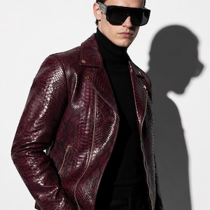 May include: A burgundy leather jacket with a snakeskin pattern, featuring a classic biker jacket design with a notched collar and a zipper closure. The jacket is paired with black sunglasses and a black turtleneck.