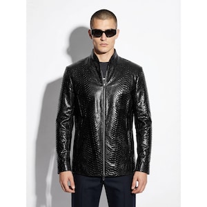 Può includere: Giacca in pelle nera con motivo strutturato e cerniera a tutta lunghezza. La giacca ha un collo alto e maniche lunghe. La persona nell'immagine indossa occhiali da sole. La giacca è probabilmente adatta a temperature più fresche.