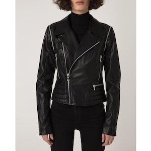 Könnte beinhalten: Schwarze Lederjacke mit asymmetrischem Reißverschluss. Die Jacke hat silberne Reißverschlüsse an den Ärmeln und Taschen, einen Reverskragen und Steppdetails an Schultern und unterem Rücken. Das Model trägt einen schwarzen Rollkragenpullover und eine schwarze Hose.