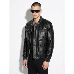 Pode incluir: Casaco bomber de couro preto com um padrão texturizado de pele de cobra. O casaco tem dois bolsos frontais, gola, punhos e bainha canelados. O modelo usa óculos de sol pretos e calças pretas.