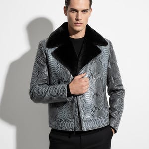 May include: A gray snakeskin-patterned jacket with a black fur collar. The jacket has a zipper closure and is worn over a black shirt. The jacket's design features a textured pattern, offering a stylish and luxurious look. The model is wearing black pants.
