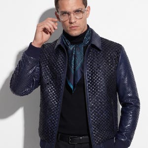 May include: A navy blue leather jacket with a woven pattern. The jacket has a collar, a zipper, and dark blue ribbed cuffs. A patterned scarf is worn under the jacket. The person is wearing glasses and a black turtleneck.