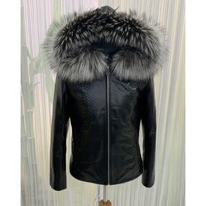 May include: A black leather jacket with a textured pattern and a silver zip. The jacket features a large hood trimmed with fluffy, grey and silver fur. The jacket is displayed on a wooden stand.