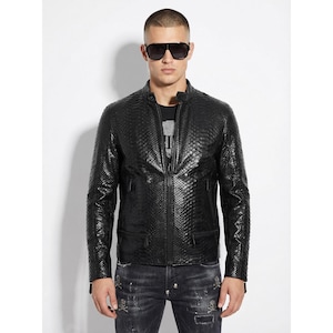 Op de afbeelding: Zwarte leren jas met een textuur, slangenleerachtig patroon. De jas heeft een ritssluiting aan de voorkant, twee zijzakken en een opstaande kraag. De persoon op de foto draagt een zonnebril en een donkerblauwe jeans.