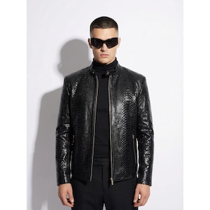 Pode incluir: Casaco de couro preto com padrão de pele de cobra texturizado. O casaco tem fecho de correr, gola alta e corte justo. O modelo usa óculos de sol pretos e uma gola alta preta.