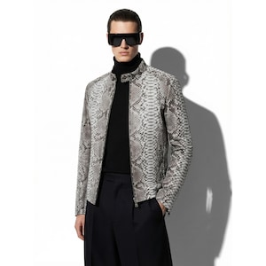 May include: A snakeskin-effect jacket in grey and white tones. The jacket features a zip closure and a stand-up collar. The person in the image is wearing a black roll neck and black sunglasses. The jacket is a stylish outerwear item.