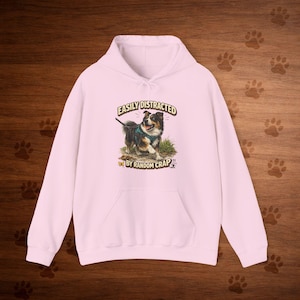 May include: A light pink hoodie with a front pocket and a drawstring hood. The front of the hoodie features a graphic of a dog on a leash with the text "EASILY DISTRACTED BY RANDOM CRAP".