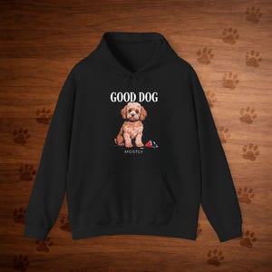 Op de afbeelding: Zwarte hoodie met een afbeelding van een lichtbruine hond met de tekst "GOOD DOG" erboven en "MOSTLY" eronder. De hond zit met een rood en zwart speeltje. De hoodie heeft een voorzak en een capuchon met een trekkoord.
