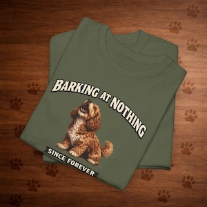 May include: Olive green t-shirt featuring a brown and white dog illustration. The text "BARKING AT NOTHING" arches above the dog, with "SINCE FOREVER" below. The shirt is folded, showing the design on the top layer.