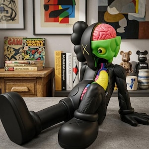 May include: A black vinyl art figure with exposed internal organs, including a pink brain and yellow lungs. The figure has a green face and a black body with an "X" on one eye. The figure is sitting with its legs extended. Books and other art pieces are in the background.