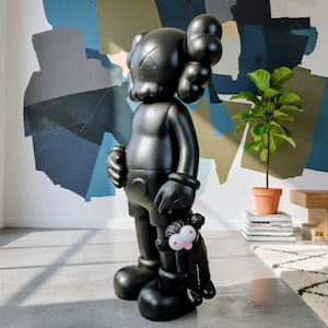 Puede incluir: Una gran escultura negra brillante de un personaje de dibujos animados con ojos tachados y una cabeza redonda, sosteniendo un peque&ntilde;o juguete de peluche negro con ojos rosados. La figura est&aacute; en una habitaci&oacute;n con un mural abstracto colorido.