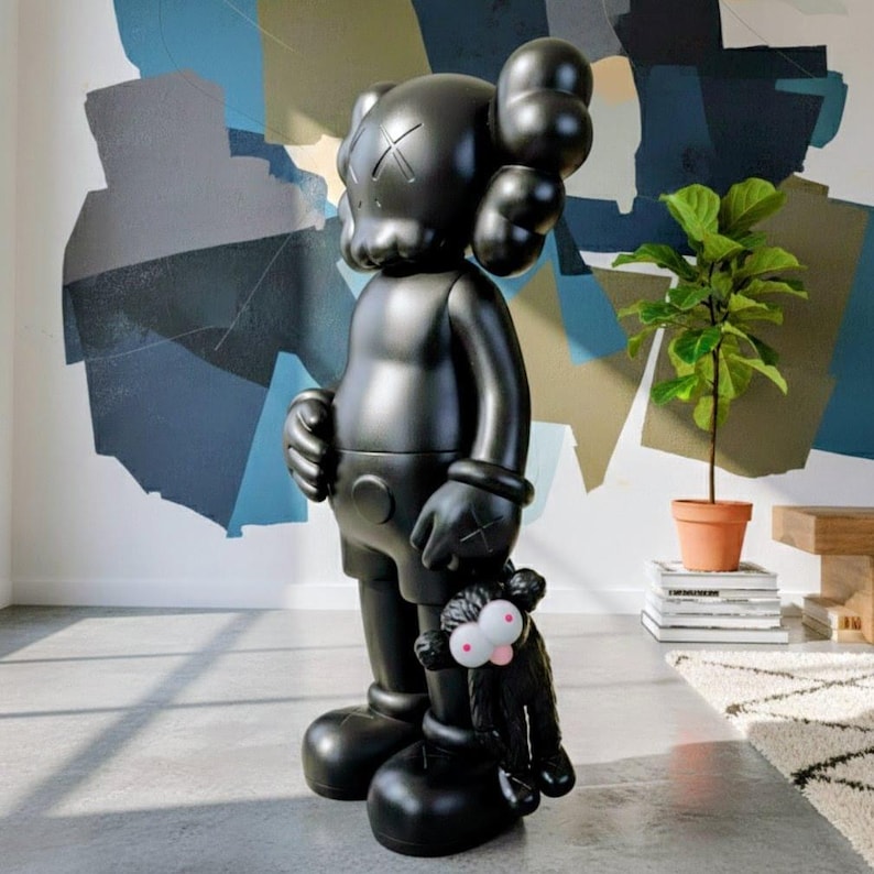 Puede incluir: Una gran escultura negra brillante de un personaje de dibujos animados con ojos tachados y una cabeza redonda, sosteniendo un peque&ntilde;o juguete de peluche negro con ojos rosados. La figura est&aacute; en una habitaci&oacute;n con un mural abstracto colorido.