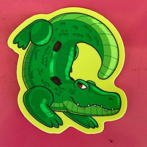 Könnte beinhalten: Ein grüner Cartoon-Alligator-Aufkleber mit gelbem Rand. Der Alligator hat ein detailliertes, strukturiertes Design mit braunen Flecken und roten Augen. Der Aufkleber befindet sich auf einer rosa Oberfläche.