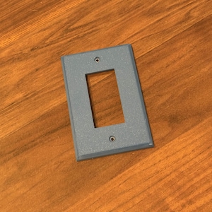 Puede incluir: Una placa de pared rectangular azul claro con una sola abertura rectangular. La placa tiene dos pequeños orificios de montaje y una superficie ligeramente texturizada. La placa se muestra sobre una superficie de madera.