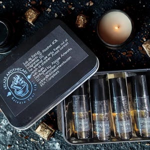 Könnte beinhalten: Eine schwarze rechteckige Metalldose mit vier kleinen Glas-Roll-on-Parfümfläschchen mit schwarzen Verschlüssen. Der Deckel trägt ein schwarzes Etikett mit dem Text Ablaze Apothecary, Release Your Light und Inhaltsstoffangaben. Die Fläschchen sind mit Almost Healed, Awakening Ritual, Nothing Faux und Ethereal Cascade beschriftet.