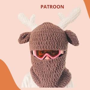 Op de afbeelding: Een bruine gehaakte skihelmhoes met witte geweien en een gezichtsbedekking. De hoes heeft een nekwarmer en wordt gedragen met een roze skibril. De tekst "RENDIER SKIHELMCOVER PATROON" staat bovenaan.