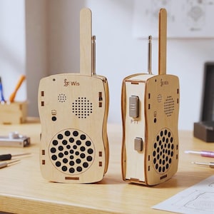 May include: Two wooden walkie-talkies with a light brown finish are displayed on a wooden desk. Each has a tall antenna, speaker holes, and the text "Wis". One has a PTT button. The design is reminiscent of vintage communication devices.