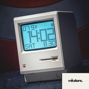 Pode incluir: Um relógio digital branco em estilo retrô com uma tela azul exibindo a hora, data e temperatura. O relógio tem um design clássico com o logotipo da Apple e uma ranhura. A hora é 14:02, sábado e 8:30. A temperatura é de 26 graus Celsius.