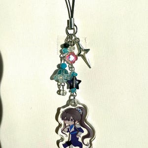 Può includere: Un ciondolo decorativo con un cordino nero, dettagli in metallo color argento e vari ciondoli. I ciondoli includono una stella, un fiore e un personaggio dei cartoni animati. Il personaggio indossa un abito blu e nero.