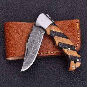 May include: A folding pocket knife with a Damascus steel blade featuring a wavy pattern. The handle is constructed from alternating diagonal segments of light brown wood and dark charcoal-coloured material, secured with small brass pins.