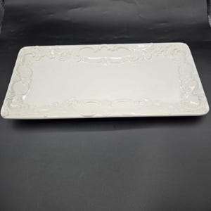 May include: A rectangular white ceramic serving tray featuring an embossed scrollwork pattern along the raised border. The center of the tray is smooth and flat, while the edges are decorated with intricate, raised relief detailing in white.