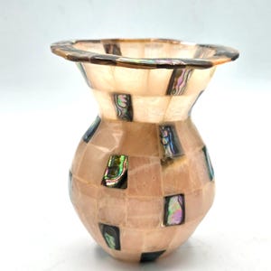 May include: A small vase featuring a mosaic pattern of light pink, translucent stone tiles interspersed with rectangular, iridescent abalone shell inlays. The vessel has a wide, flared rim and a rounded body, showcasing a smooth, polished finish.