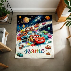 Puede incluir: Una colorida manta con coches de dibujos animados corriendo en una pista en el espacio. El dise&ntilde;o incluye planetas, un sat&eacute;lite y la palabra "name" en cursiva. Los coches son de varios colores, con un vibrante salpicado de arco&iacute;ris.