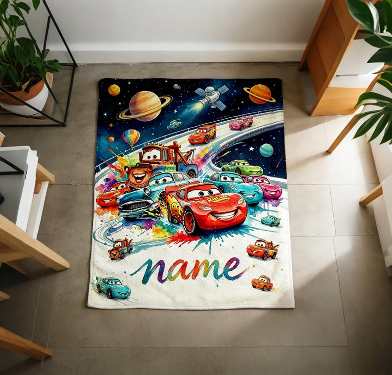 Puede incluir: Una colorida manta con coches de dibujos animados corriendo en una pista en el espacio. El dise&ntilde;o incluye planetas, un sat&eacute;lite y la palabra "name" en cursiva. Los coches son de varios colores, con un vibrante salpicado de arco&iacute;ris.