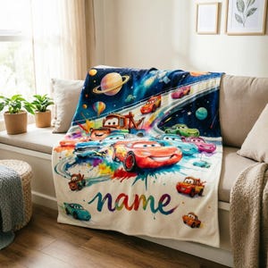 Puede incluir: Una manta de color crema con un dise&ntilde;o colorido de coches de carreras de dibujos animados en un entorno espacial. La manta tiene un nombre personalizado en un estilo de acuarela, con varios coches corriendo en una pista.