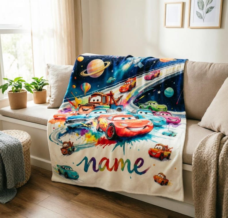 Puede incluir: Una manta de color crema con un dise&ntilde;o colorido de coches de carreras de dibujos animados en un entorno espacial. La manta tiene un nombre personalizado en un estilo de acuarela, con varios coches corriendo en una pista.