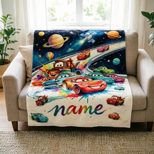Puede incluir: Una colorida manta con tem&aacute;tica de Cars que presenta una escena espacial con planetas, un sat&eacute;lite y coches de carreras. La manta tiene una base blanca con la palabra "name" en escritura cursiva. La manta est&aacute; sobre un sof&aacute; beige.