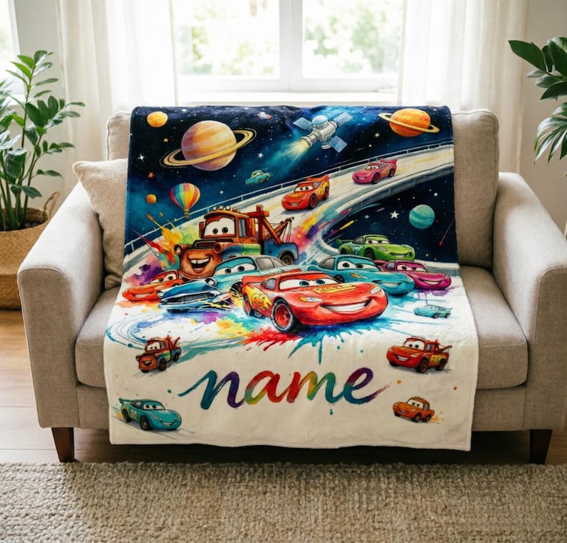 Puede incluir: Una colorida manta con tem&aacute;tica de Cars que presenta una escena espacial con planetas, un sat&eacute;lite y coches de carreras. La manta tiene una base blanca con la palabra "name" en escritura cursiva. La manta est&aacute; sobre un sof&aacute; beige.
