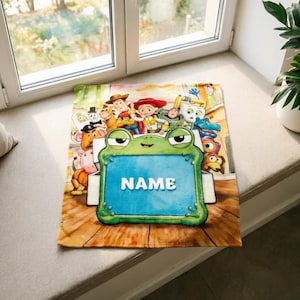 May include: A rectangular blanket featuring a colorful illustration of various animated characters from the Toy Story franchise surrounding a large, central green frog-shaped frame with the word NAME printed in white capital letters.