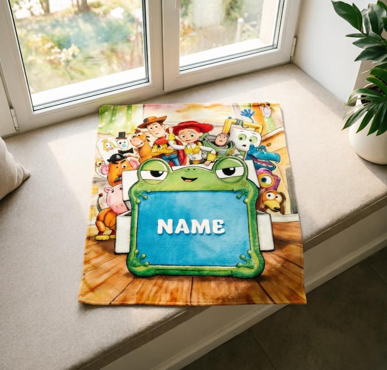 May include: A rectangular blanket featuring a colorful illustration of various animated characters from the Toy Story franchise surrounding a large, central green frog-shaped frame with the word NAME printed in white capital letters.