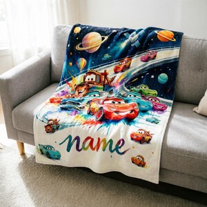 Puede incluir: Una manta blanca con una colorida ilustraci&oacute;n de coches de dibujos animados en el espacio. El dise&ntilde;o incluye planetas, estrellas y la palabra "name" en colores arco&iacute;ris. La manta est&aacute; sobre un sof&aacute; gris.