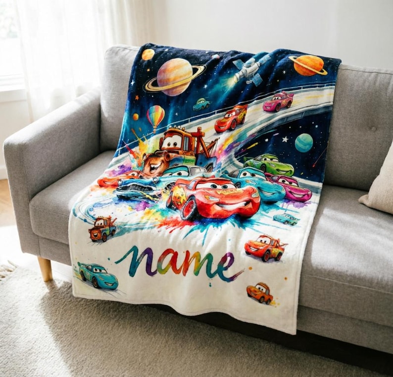 Puede incluir: Una manta blanca con una colorida ilustraci&oacute;n de coches de dibujos animados en el espacio. El dise&ntilde;o incluye planetas, estrellas y la palabra "name" en colores arco&iacute;ris. La manta est&aacute; sobre un sof&aacute; gris.