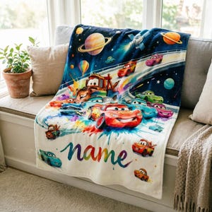 Puede incluir: Una manta blanca y suave con una colorida ilustraci&oacute;n de coches de carreras en un entorno espacial. La manta tiene la palabra "name" en una fuente de arco&iacute;ris. La manta est&aacute; sobre un sof&aacute; gris.