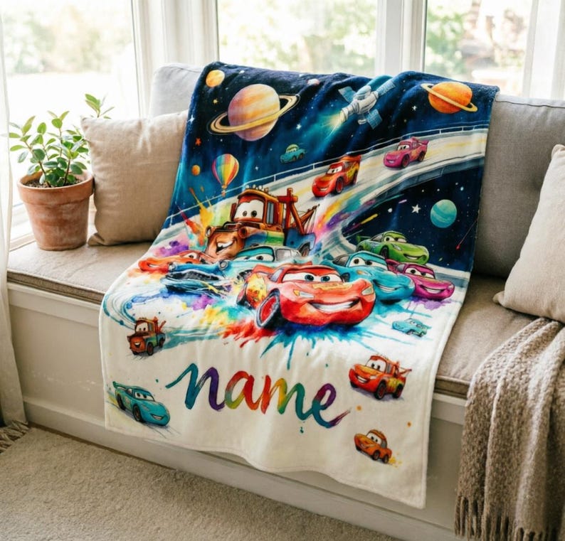 Puede incluir: Una manta blanca y suave con una colorida ilustraci&oacute;n de coches de carreras en un entorno espacial. La manta tiene la palabra "name" en una fuente de arco&iacute;ris. La manta est&aacute; sobre un sof&aacute; gris.