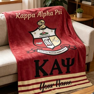 May include: A maroon blanket with the text "Kappa Alpha Psi" in gold script. The blanket features a crest with a knight's helmet and the letters "KAY" in black. The bottom has a cream-colored stripe with the words "Your Name".