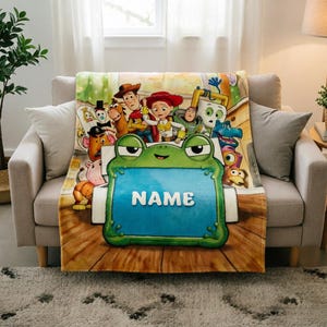 May include: A soft throw blanket featuring a colorful graphic print of various animated characters from the Toy Story franchise, including Woody, Jessie, and Buzz Lightyear, centered around a large green frog holding a blue sign with the word NAME.