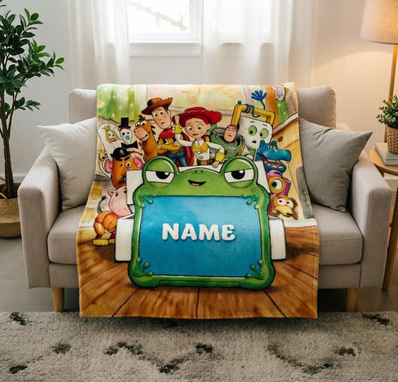 May include: A soft throw blanket featuring a colorful graphic print of various animated characters from the Toy Story franchise, including Woody, Jessie, and Buzz Lightyear, centered around a large green frog holding a blue sign with the word NAME.