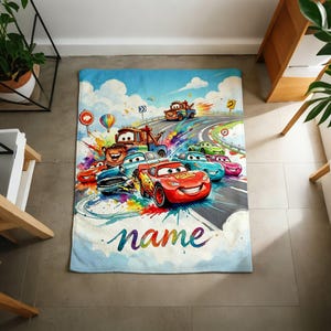 Puede incluir: Una colorida manta con tem&aacute;tica de Cars que presenta a Rayo McQueen y otros personajes corriendo en una pista. El dise&ntilde;o incluye un cielo azul, una carretera y la palabra "name" en colores arco&iacute;ris. La manta es rectangular.