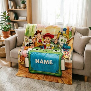 May include: A soft fleece blanket featuring a colorful illustration of various animated toy characters, including a cowboy, a cowgirl, and a space ranger, gathered behind a large, green, frog-shaped toy box with the word NAME printed in white.