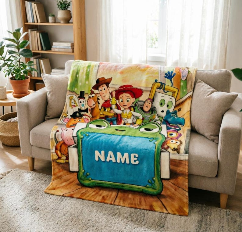 May include: A soft fleece blanket featuring a colorful illustration of various animated toy characters, including a cowboy, a cowgirl, and a space ranger, gathered behind a large, green, frog-shaped toy box with the word NAME printed in white.