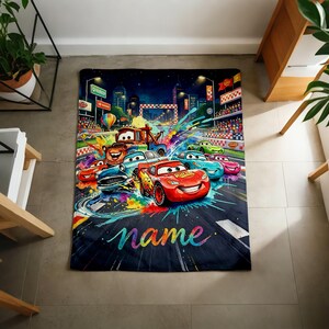 Puede incluir: Una colorida manta con coches de dibujos animados corriendo en una pista. El dise&ntilde;o incluye un fondo de paisaje urbano, una l&iacute;nea de meta y la palabra "name" en una fuente de arco&iacute;ris. La manta es una vibrante mezcla de colores.